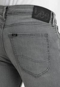 Šedé džíny z denimu s texturovanou tkaninou, se štítkem z černé kůže na pase s embosovaným nápisem "Lee" a zadními kapsami se jednoduchým prošíváním.