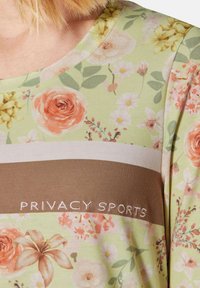 Hellgrünes Oberteil mit floralem Druck, der pinke und gelbe Blumen zeigt. Brauner horizontaler Streifen mit "PRIVACY SPORTS" in Weiß gestickt.