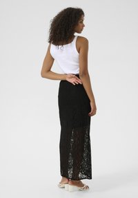 Jupe maxi en dentelle noire avec des panneaux transparents, complétée par un débardeur blanc ajusté. Des cheveux bouclés et des chaussures minimalistes achèvent le look.