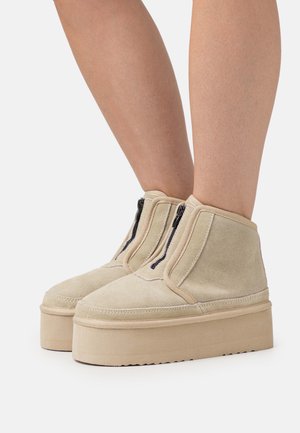 Bottines en suède beige avec fermetures éclair noires, dotées d'une épaisse semelle plateforme et d'un bout rond. Texture lisse, coutures minimales.