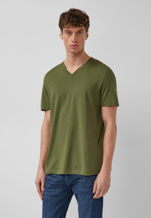 Mężczyzna ubrany w oliwkowozieloną koszulkę z krótkim rękawem i dekoltem w serek oraz niebieskie dżinsy, stojący na tle jednolitego, jasnoszarego tła.