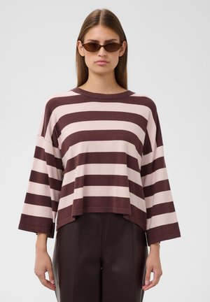 Femme aux longs cheveux portant des lunettes de soleil marron, un pull ample rayé rose et marron, et un pantalon marron foncé sur un fond uni.