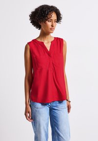 Czerwona bluzka bez rękawów z dekoltem w serek, dwoma kieszeniami na piersi i lekko luźnym krojem, wykonana z gładkiego materiału. Połączona z jasnoniebieskimi dżinsami.