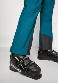 Black ski boots with green accents feature adjustable metal buckles. Teal ski pants with black reinforced cuffs are shown from knee to ankle.