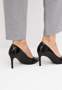 Zapatos de tacón alto de charol negro con punta afilada y un tacón stiletto delgado, combinados con pantalones grises claros ajustados.