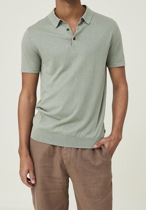 Hellgrünes Polohemd mit kurzen Ärmeln, Kragen und drei Knöpfen, kombiniert mit braunen Leinenhosen in lässiger Passform.