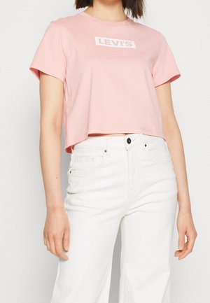 Vrouw draagt een cropped lichtroze Levi's T-shirt en hoog getailleerde witte jeans, staand tegen een effen lichte achtergrond.
