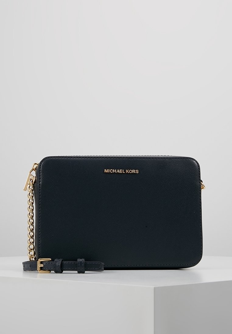 Sac en bandoulière rectangulaire noir de Michael Kors avec une chaîne dorée et un logo à l'avant, reposant sur une surface blanche contre un fond gris.