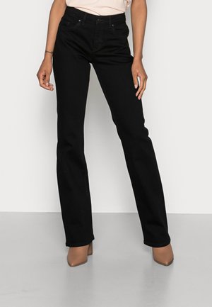 Mujer con jeans negros de corte recto y tacones beige con punta afilada, de pie frente a un fondo claro y liso.