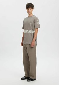 Grå bomulls-T-skjorte med korte ermer, med hvit tekst og logoer, kombinert med beige vide bukser og svarte sko.