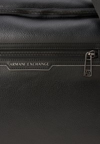 Crna kožna torba s vidljivom teksturom, zatvaranje na munju i označena oznakom "Armani Exchange". Ima metalni zatvarač s dekorativnom ručkom.