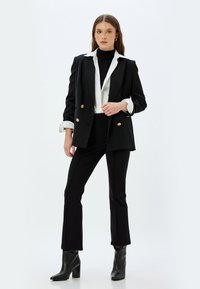 Musta kaksirivinen puku, jossa on kultanappi, valkoisen paidan ja mustan poolopaidan päällä, yhdistettynä kapeisiin mustiin housuihin ja nilkkureihin.