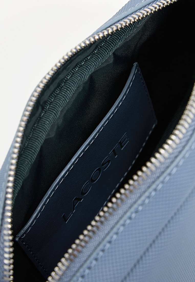 Intérieur d'un sac synthétique texturé bleu marine avec une doublure verte et une étiquette Lacoste visible. Comprend des détails de fermeture éclair en argent.