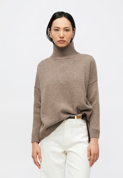 Femme portant un pull à col roulé côtelé marron rentré dans un pantalon blanc taille haute avec une ceinture foncée à boucle dorée, debout devant un fond uni.