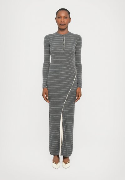 Robe maxi à rayures grises avec des manches longues, un col rond, une fermeture à boutons sur le devant et un panneau blanc asymétrique. Portée avec des chaussures blanches.