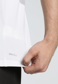 Valge sportlik särk, millel on tekstuuriga disain, kerge materjal ja nähtav "DRYCELL" silt helmel. Kohandatud ja sobib sportimiseks.