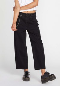 Zwarte wijde spijkerbroek van denim, met een kettingdetail op de tailleband, en gecombineerd met glanzende zwarte schoenen.
