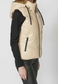 Femme portant une veste sans manches matelassée beige à capuche avec cordons noirs par-dessus un top noir à manches longues et un pantalon noir, vue de côté.