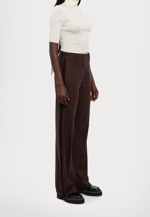 Personne portant un col roulé blanc à manches courtes, un pantalon marron à jambes larges avec des détails sur les côtés, et des chaussures noires à semelle épaisse, debout sur un fond blanc.