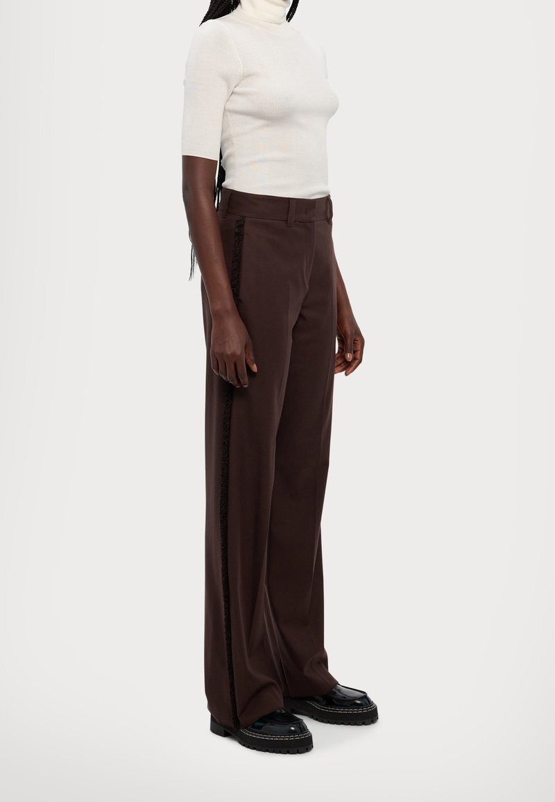 Personne portant un col roulé blanc à manches courtes, un pantalon marron à jambes larges avec des détails sur les côtés, et des chaussures noires à semelle épaisse, debout sur un fond blanc.