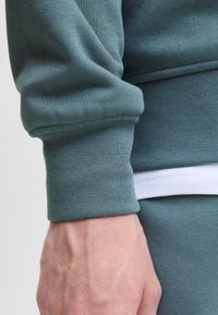 Groene sweatshirtmanchet met een textuurstof. Witte tailleband zichtbaar onder de manchet, waardoor het contrast met het materiaal wordt benadrukt.