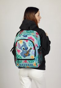 Sac à dos avec une base turquoise ornée de motifs floraux colorés et d'un personnage de dessin animé jouant de la guitare. Comprend des poches latérales en maille noire.