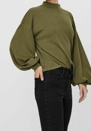 Person trägt einen olivgrünen, strukturierten Pullover mit voluminösen Ärmeln und schwarze hoch taillierte Jeans, Hand in der Tasche, vor einfachem Hintergrund.