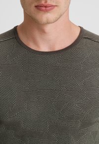 T-shirt en maille texturée vert olive présentant un motif géométrique subtil, un col rond et un tissu de poids moyen.