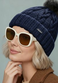 Navy knitted beanie with a pom-pom, paired with oversized white sunglasses featuring brown tinted lenses, set against a light background.