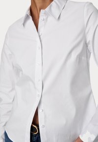 Biała koszula z długim rękawem, wykonana z gładkiej bawełny, z klasycznym kołnierzykiem, z przodu zapinana na guziki oraz z pojedynczą kieszonką na piersi. Wytrzymałe szycie.