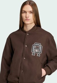 Brun bomberjakke med trykknapper, ribbet krave og hvidt grafisk logo med "Adidas Originals" og et stiliseret design.