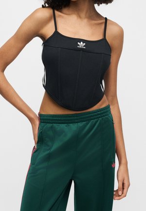 Kobieta ubrana w czarny krótki top Adidas z cienkimi ramiączkami i zielone spodnie sportowe Adidas z czerwonymi paskami.