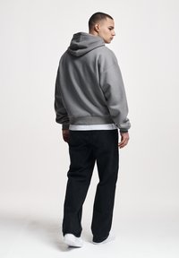 Joven de perfil vistiendo una sudadera con capucha gris, camiseta blanca, vaqueros negros y zapatillas blancas sobre un fondo blanco liso.