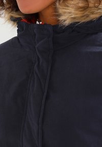 Námořnická modrá izolovaná bunda s kapucí lemovanou umělou kožešinou, se zipem a knoflíkovým uzávěrem a prošívanou texturou na přední straně.