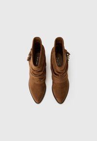Brown suede ankle boots with a slouch design, pointed toe, and decorative fringe detail on the side. Smooth texture with a flat sole.