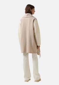 Pletená vesta z světle béžové vlny, vrstvená přes krémovou prošívanou bundu se šálovým límcem a třásněmi na lemu. Bílé kalhoty a béžové boty.