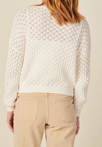 White knit sweater with a textured open weave and ribbed cuffs, paired with light beige jeans featuring two back pockets and subtle stitching details.
