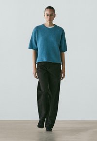 Giovane donna con capelli raccolti che indossa un maglione blu a maniche corte, pantaloni neri a gamba larga e scarpe nere, che cammina in avanti su un pavimento chiaro.