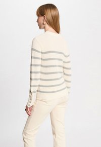 Pull en tricot rayé crème avec des rayures horizontales gris clair, poignets côtelés et détails décoratifs aux bords des manches, associé à un pantalon beige.