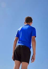 Modra športna majica s kratkimi rokavi in gladko teksturo, kombinirana s črnimi kratkimi hlačami. Ozadje je jasno modro nebo.