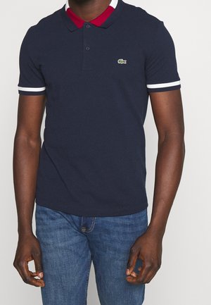 Mann trägt ein marineblaues Poloshirt mit rotem Innenlayer und weißem Kragen sowie weißen gestreiften Ärmelbündchen, kombiniert mit blauen Jeans.