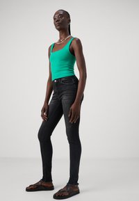Zelen majica na naramenice, črne skinny jeans z izbledelim izgledom in črne sandale s prečnimi naramnicami. Model stoji v profilu.