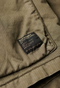 Tissu en vert olive à motif en chevrons avec une étiquette noire comportant un texte doré, détaillant la marque et les instructions d'entretien, cousu sur le vêtement.