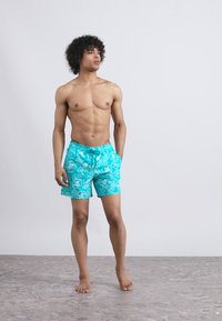 Mladenič s kodrastimi lasmi, ki stoji bos v turkiznih kopalkah z belimi tropskimi vzorci, gleda na stran proti enobarvni ozadju.