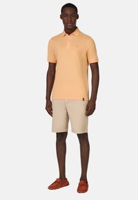 Polo triko v světle oranžové bavlně s dvouknoflíkovou légou, spárované s béžovými kraťasy a hnědými slip-on botami.