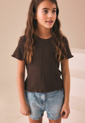 Mlada djevojka s dugom valovitom kosom, nosi smeđu kratku majicu s teksturom i svijetloplave traper kratke hlače, stoji ispred neutralne pozadine.