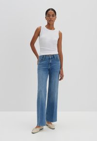 Canottiera bianca a coste, jeans a gamba larga blu chiaro e scarpe basse beige. La modella posa contro uno sfondo bianco uniforme.