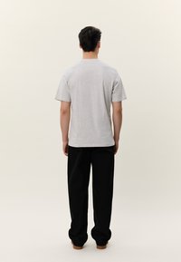 Muž stojí chrbtom k nám, má na sebe obyčajné svetlosivé tričko s krátkym rukávom, voľné čierne nohavice a hnedé topánky na svetlom pozadí.