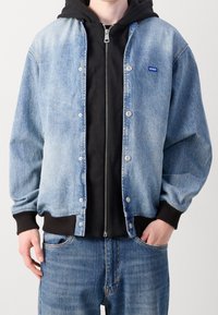 Denim jacket in light blue with button closures, ribbed cuffs, and waistband. It layers over a black zip-up hoodie, creating a contrasting look.