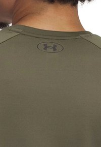 Camiseta deportiva verde oliva con un logotipo negro redondo en la parte superior de la espalda, con textura de tela suave y mangas raglán.